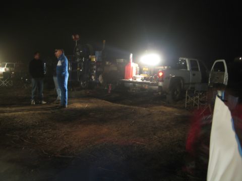 1990 Ford Bronco - Two men stand near a lit pit area with fuel jugs and trucks on a dark desert lot at night.