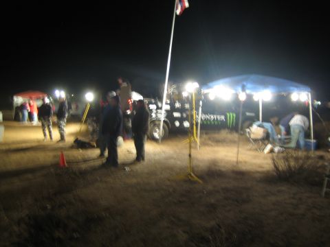 1990 Ford Bronco - People gather around floodlit pit canopies, including a Monster Energy tent, on dark desert scrubland at night.