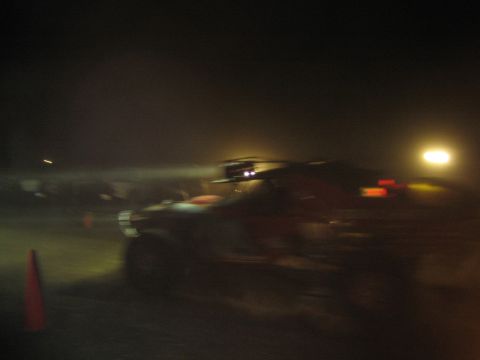 1990 Ford Bronco - blurred off-road race truck with roof lights speeds past an orange cone in a dusty pit at night.
