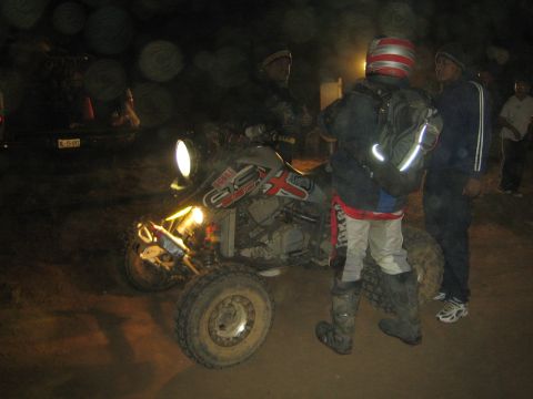 1990 Ford Bronco - rider in a striped helmet and backpack stands beside a quad ATV talking with others on a dirt lot at night.