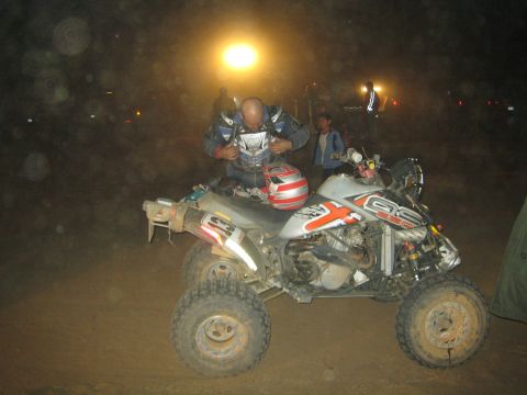 1990 Ford Bronco - rider in protective gear stands over a silver sport quad ATV marked with an X, with a red helmet on the seat, at a dimly lit night off-road…
