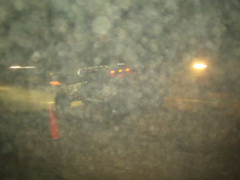 1990 Ford Bronco - dark off-road race vehicle with marker lights drives past at night, viewed through a rain-spotted, out-of-focus foreground.