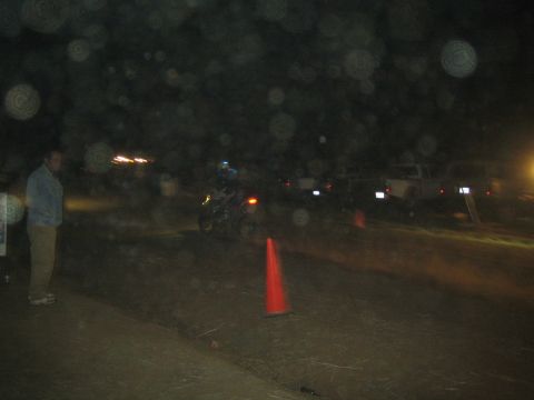 1990 Ford Bronco - dirt bike rider rides past orange traffic cones on a dark dirt course as a man and parked trucks watch from the side.