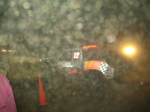 1990 Ford Bronco - off-road race truck numbered 12 with a Collins Motorsports decal and checkered-flag graphic drives past traffic cones at night.