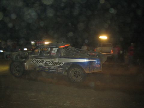1990 Ford Bronco - blue and white off-road race truck with Pro Comp lettering and number 5 stirs up dust at a night off-road event with spectators.