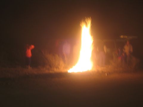 1990 Ford Bronco - tall bonfire flame burns brightly at night as silhouetted people and a vehicle stand around it on open ground.