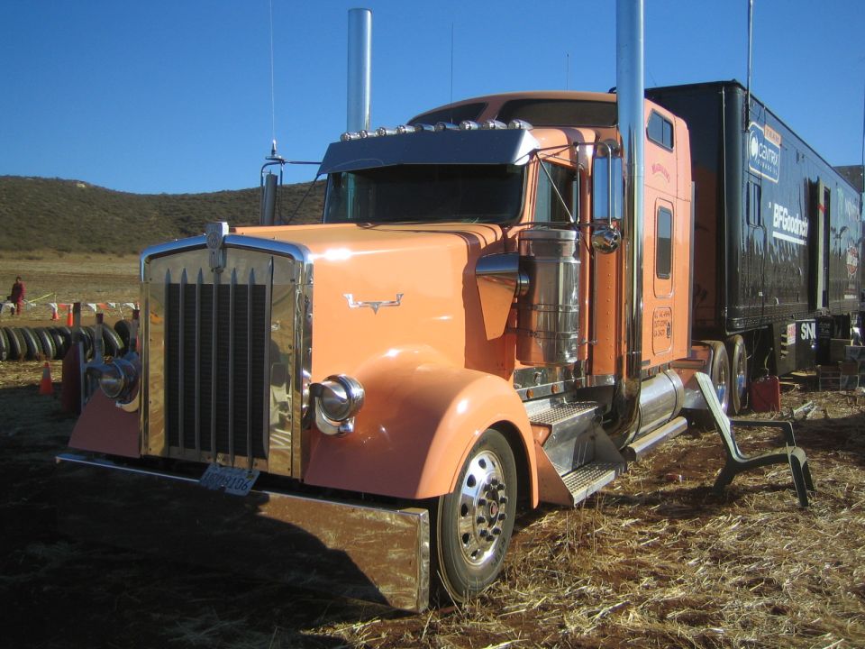 Orange long-nose semi truck with chrome grille parked in a dirt field