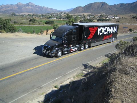 1990 Ford Bronco - black Volvo semi truck hauling a Yokohama Motorsports trailer travels a two-lane road through a valley framed by mountains.