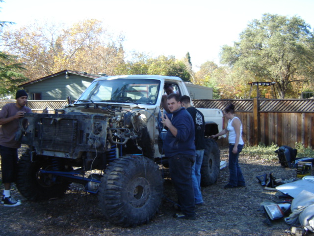 Stripped white truck/SUV on big mud tires being torn down in a backyard, people standing around it