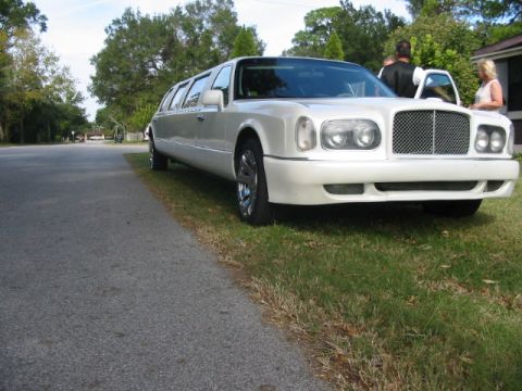 1997 Ford Crown Victoria - A long white limousine is parked on the side of a road, with people standing near the open front passenger door.