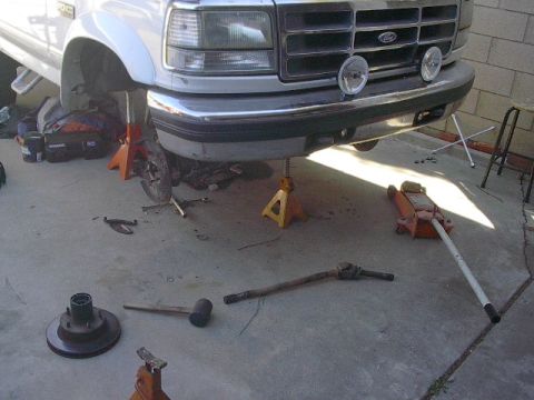1993 Ford Bronco - Front end of a white 1993 Ford Bronco is raised on jack stands, with various parts scattered on the ground.