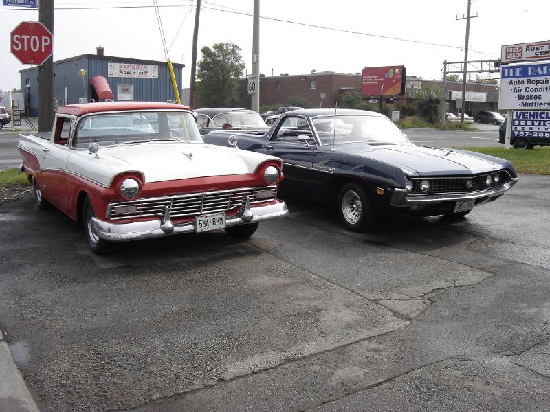 Red-and-white 1957 Ford Ranchero beside a dark Ford Torino at a stop sign and auto shop