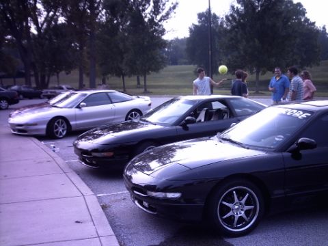 1995 Ford Probe - A group of people gather near several Ford Probes parked in a lot on a cloudy day.