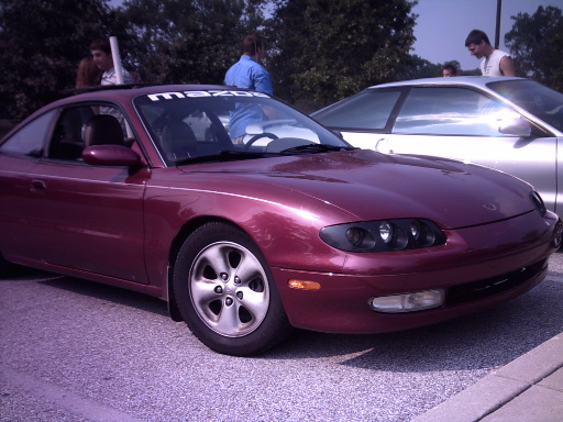 Maroon Mazda MX-6 coupe parked next to a silver car, with 'mazda' banner on windshield