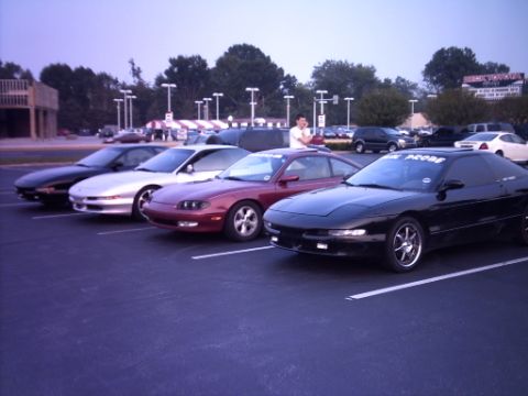 1995 Ford Probe - A line of Ford Probes are parked in a lot, with the closest one being black and having text on its windshield.