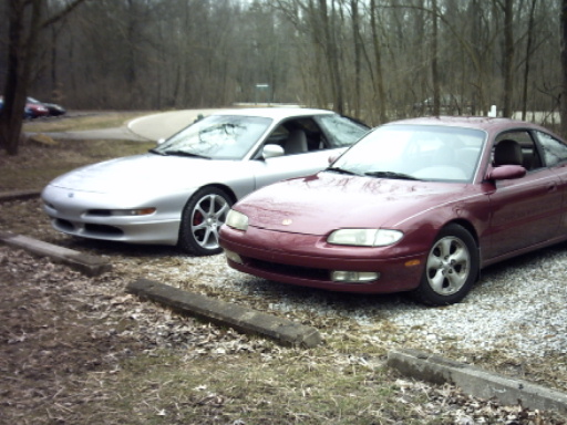 Maroon and silver coupes parked on gravel lot with trees in background. "SUPERMOTORS" logo visible