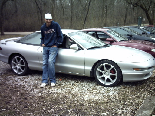 Man in blue hoodie and jeans leaning on a silver 1995 Ford Probe; other cars in background