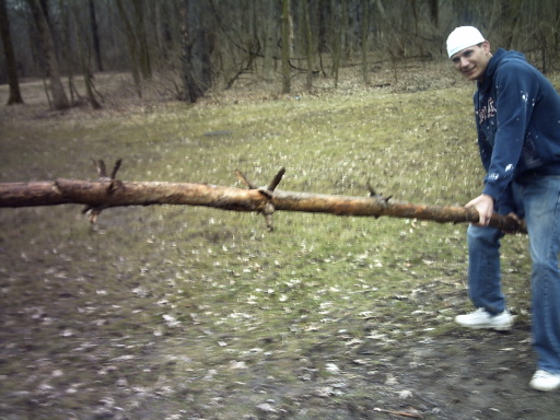 guy in a white hat and blue hoodie holding a long log in a wooded area