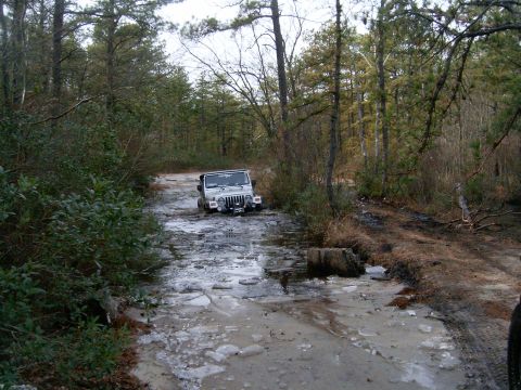 1986 Ford F-250 - A silver Jeep Wrangler ventures through a challenging muddy and icy path in a wooded area.