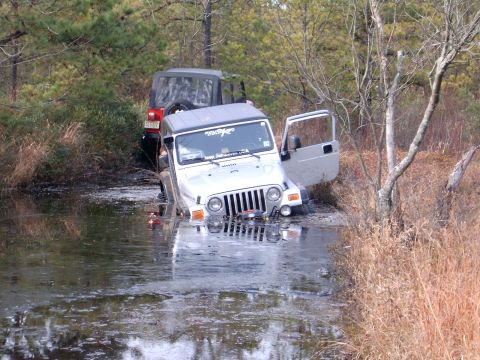 1986 Ford F-250 - Two Jeeps are stuck in the water. The front Jeep is a silver Wrangler with its door open.
