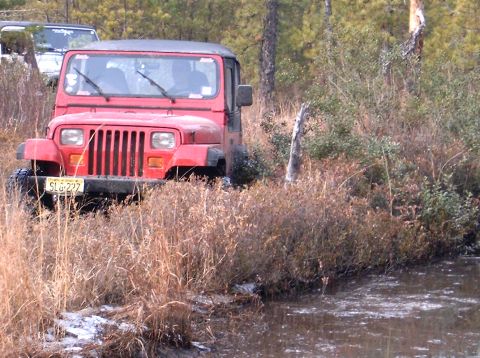1986 Ford F-250 - A red Jeep Wrangler navigates through a muddy, wooded area with another vehicle visible in the background.