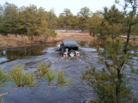 1986 Ford F-250 - A Jeep Wrangler navigates through a water crossing surrounded by trees and dry brush.
