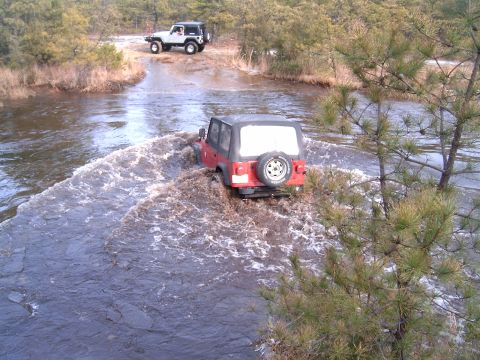 1986 Ford F-250 - Two Jeep Wranglers navigate a shallow river crossing surrounded by trees and brush.