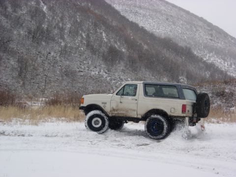 1987 Ford Bronco - A white Ford Bronco kicks up snow as it drives through a snowy landscape with a mountain in the background.