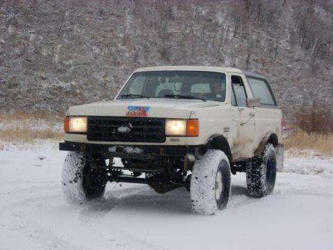 1987 Ford Bronco - A white 1987 Ford Bronco navigates a snowy terrain, its large tires kicking up snow.