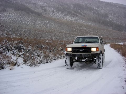 1987 Ford Bronco - A 1987 Ford Bronco navigates a snow-covered road, its tires kicking up snow as it heads towards a distant mountain.
