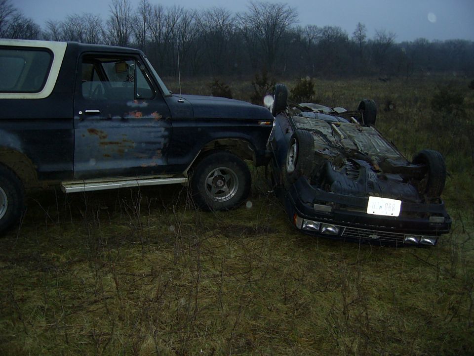 Dark blue Ford Bronco in a field beside a flipped-over car lying on its roof at dusk