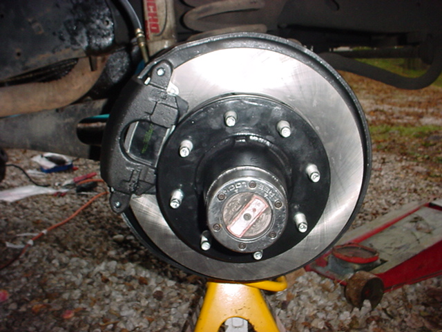Front disc brake rotor, black caliper, and manual locking hub with wheel removed, vehicle on a jack stand over gravel
