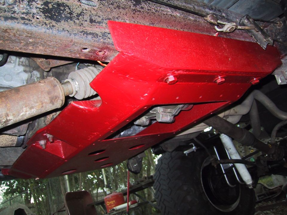 Underside view of a red skid plate bolted to a vehicle frame, showing drivetrain components and suspension
