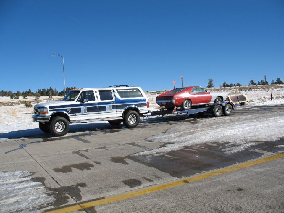 White and blue four-door Ford SUV towing a red fastback coupe on an open trailer in snow