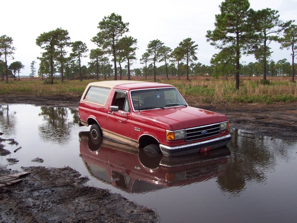 Red Ford Bronco stuck in a muddy puddle, front end submerged, tan top, side view