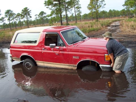 1989 Ford Bronco - A red SUV is stuck in a muddy puddle with a person attempting to push it out.