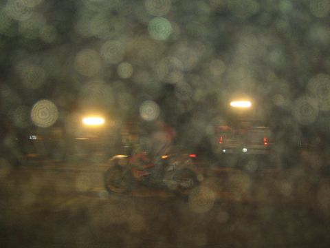 1990 Ford Bronco - motorcycle rider passes a parked pickup truck at a night off-road event, seen through a rain-spotted, out-of-focus foreground.