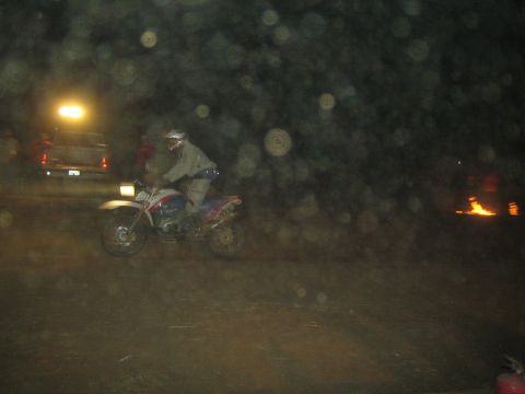 1990 Ford Bronco - helmeted rider on a red and white dual-sport dirt bike rides past parked trucks and a small fire at a dark off-road event.