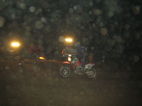 1990 Ford Bronco - helmeted rider on a red dirt bike numbered 263 rides past parked trucks and a small fire at a night off-road event.
