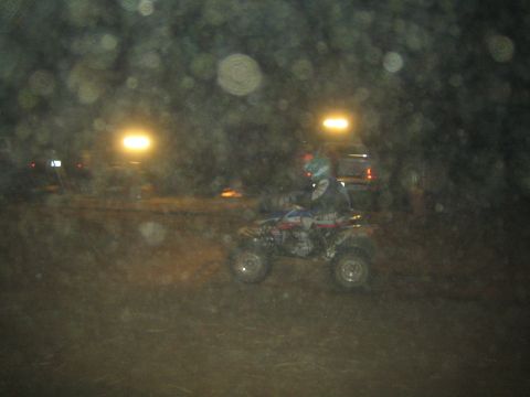 1990 Ford Bronco - helmeted rider on a sport quad ATV rides past parked trucks at a dark night off-road event, seen through a hazy foreground.