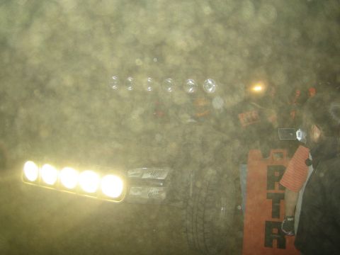 1990 Ford Bronco - off-road vehicle's five-light bar glares through thick dust at night as spectators and a person in an orange jacket stand nearby.