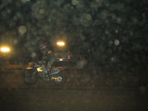 1990 Ford Bronco - helmeted rider on a dirt bike numbered 253 rides past a parked pickup truck at a night off-road event amid floating dust specks.