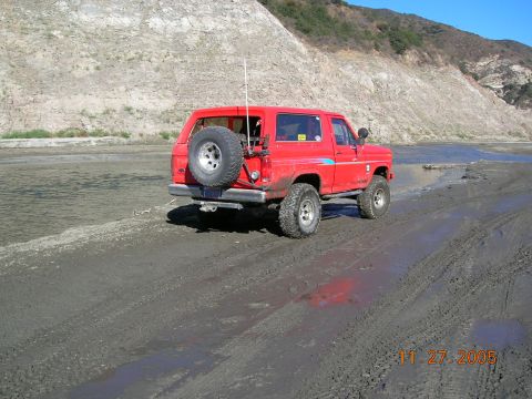 1985 Ford Bronco - Red 4x4 off-road vehicle with spare tire on rear, driving on muddy terrain near water.
