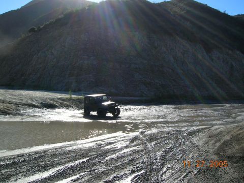 1985 Ford Bronco - A Jeep Wrangler navigates a muddy terrain under a bright sun with lens flare.