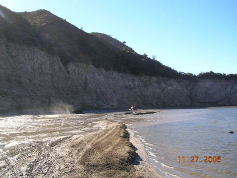 1985 Ford Bronco - Off-road vehicles navigate a challenging terrain of mud and water, with a prominent cliff face in the background.