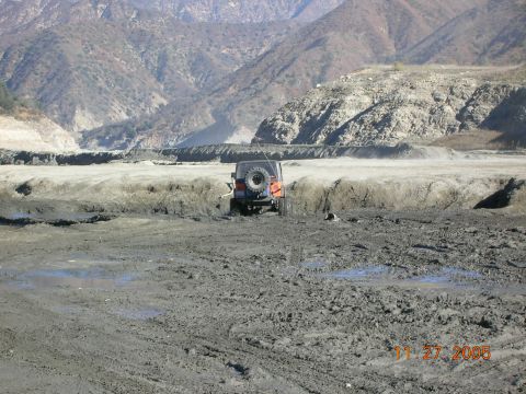 1985 Ford Bronco - Jeep Wrangler navigates a muddy terrain with mountains in the background.