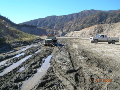 1985 Ford Bronco - Two trucks are stuck in mud on a construction site, with a person attempting to free one of them.