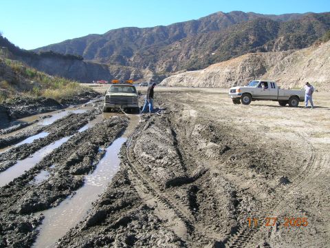 1985 Ford Bronco - Two trucks are stuck in a muddy, rutted area with mountains in the distance. One truck appears to be winching the other.