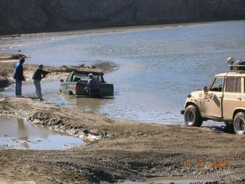 1985 Ford Bronco - Two off-road vehicles are stuck in muddy water, with onlookers observing the situation.