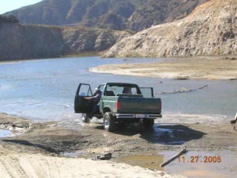 1985 Ford Bronco - A green pickup truck is partially submerged in shallow water, with its doors open. The scene is a sandy riverbank with hills in the backgro…